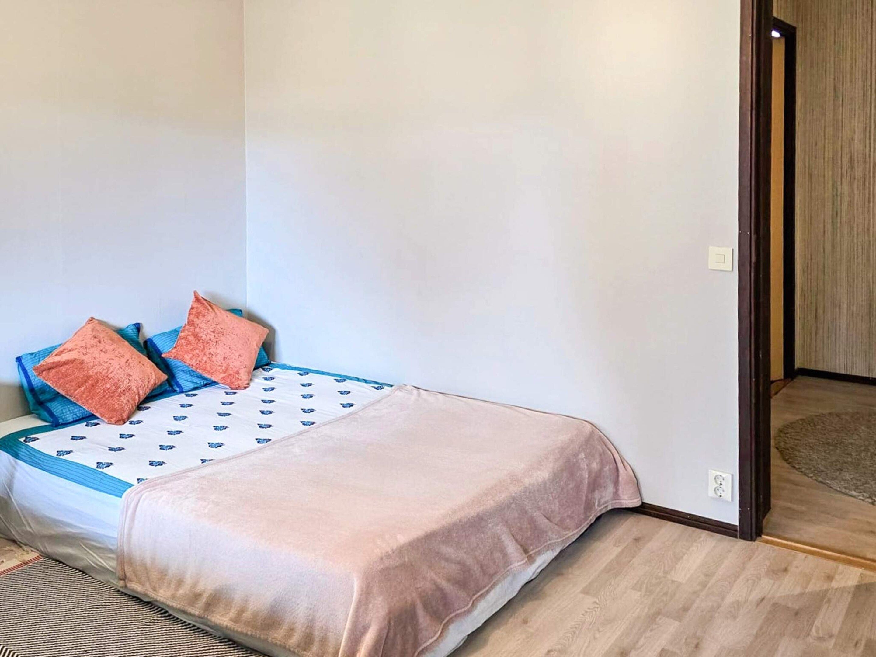 A single bed with a patterned bedspread, two pillows, and a light-colored blanket in a room with wooden flooring and a plain wall.