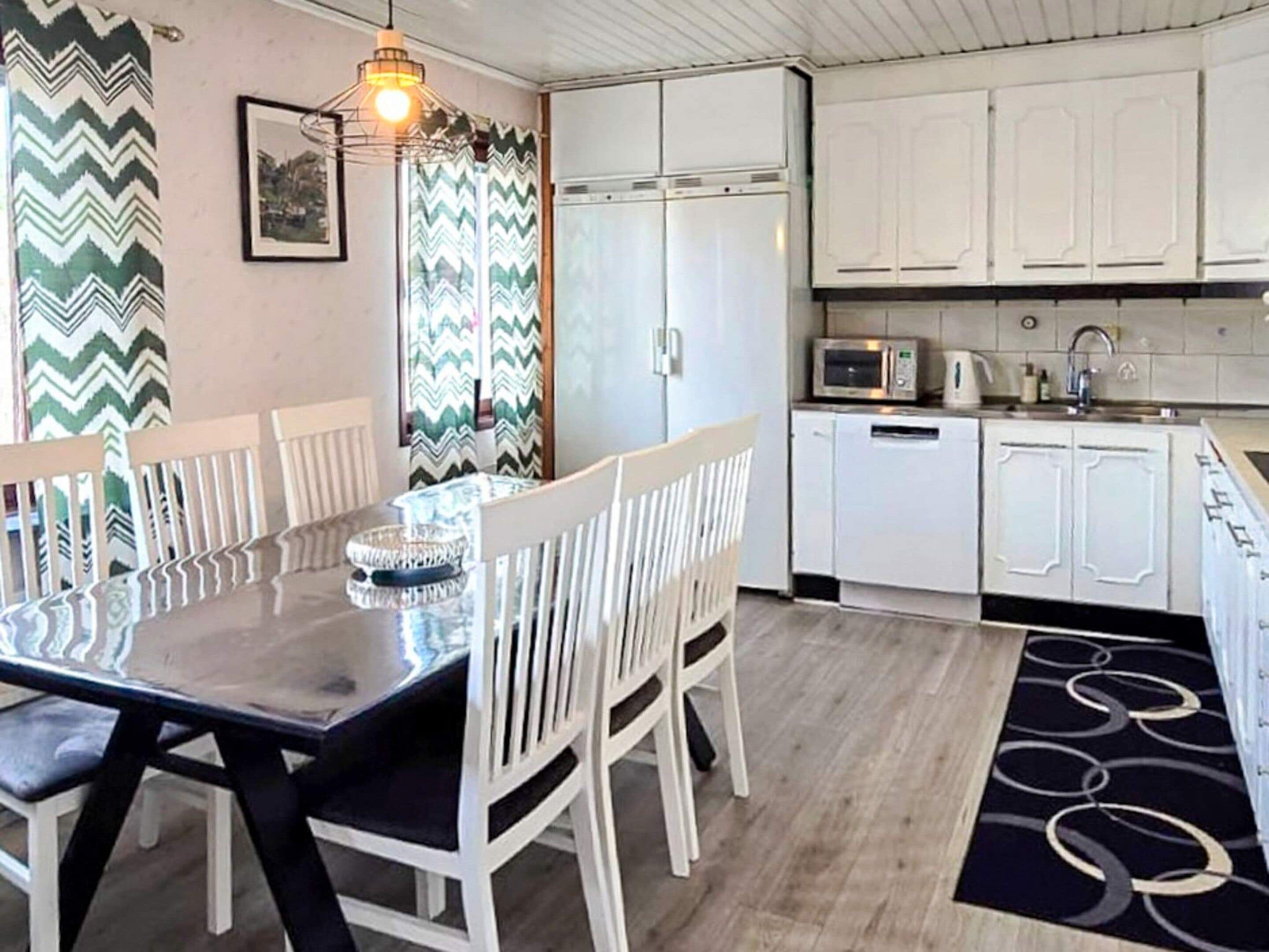 A dining area with a table and chairs, a kitchen with white cabinets and appliances, and a patterned rug.