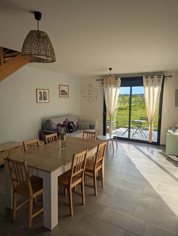 A dining area with a wooden table and chairs, a sofa, and a view of a green landscape through large windows.