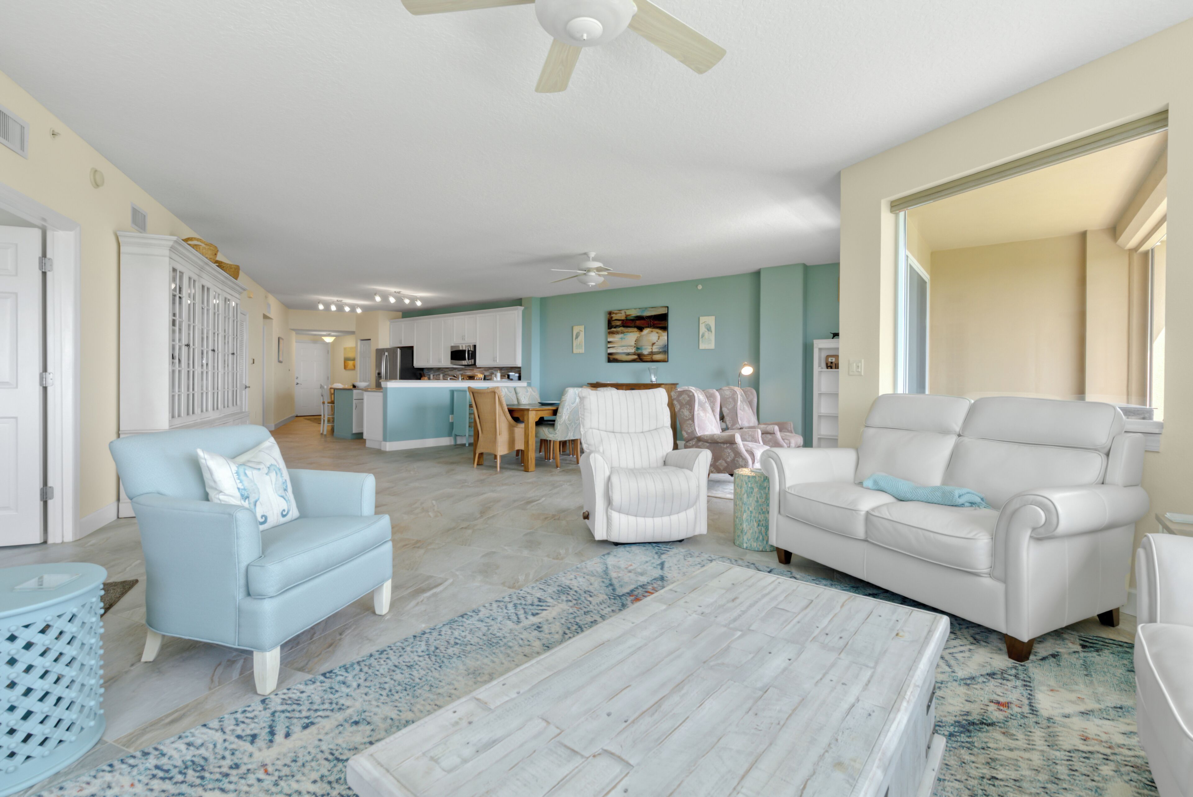 A spacious living room with a white sofa, a blue armchair, and a coffee table. There is a dining area with a table and chairs in the background.