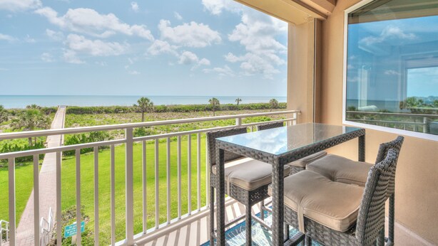 A balcony with a glass table and wicker chairs, overlooking a grassy area and the ocean.