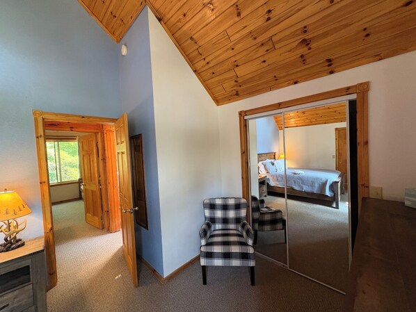 A bedroom with a wooden ceiling, a bed, a mirror, and a plaid chair.