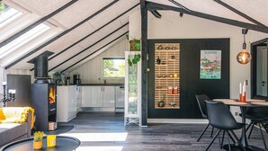 Un loft moderne avec de hauts plafonds, des poutres apparentes, une cuisine dotée d’un poêle à bois, un coin repas avec des chaises noires et une vue sur l’extérieur.