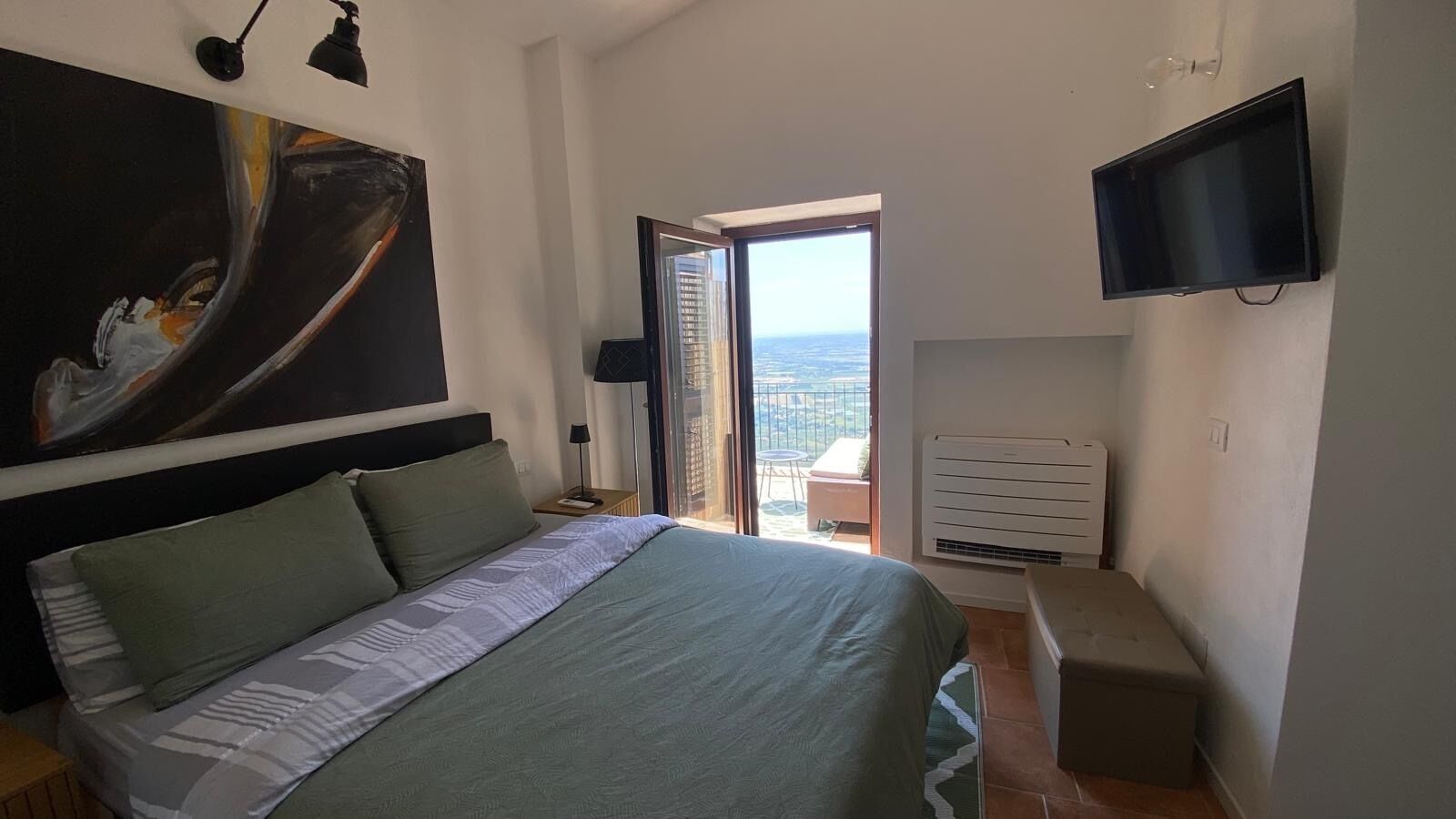 A bedroom with a large bed, a flat-screen TV, a painting, and a balcony with a view.
