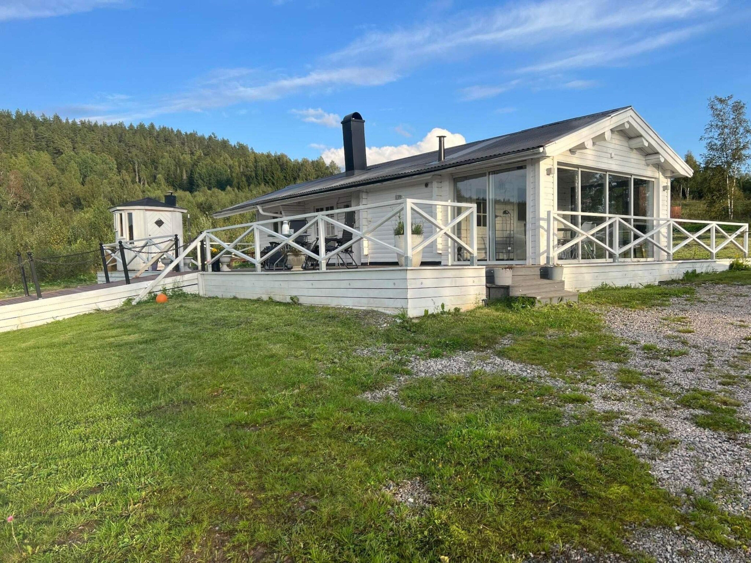 A white cabin with a covered patio, surrounded by a gravel path and grassy area.