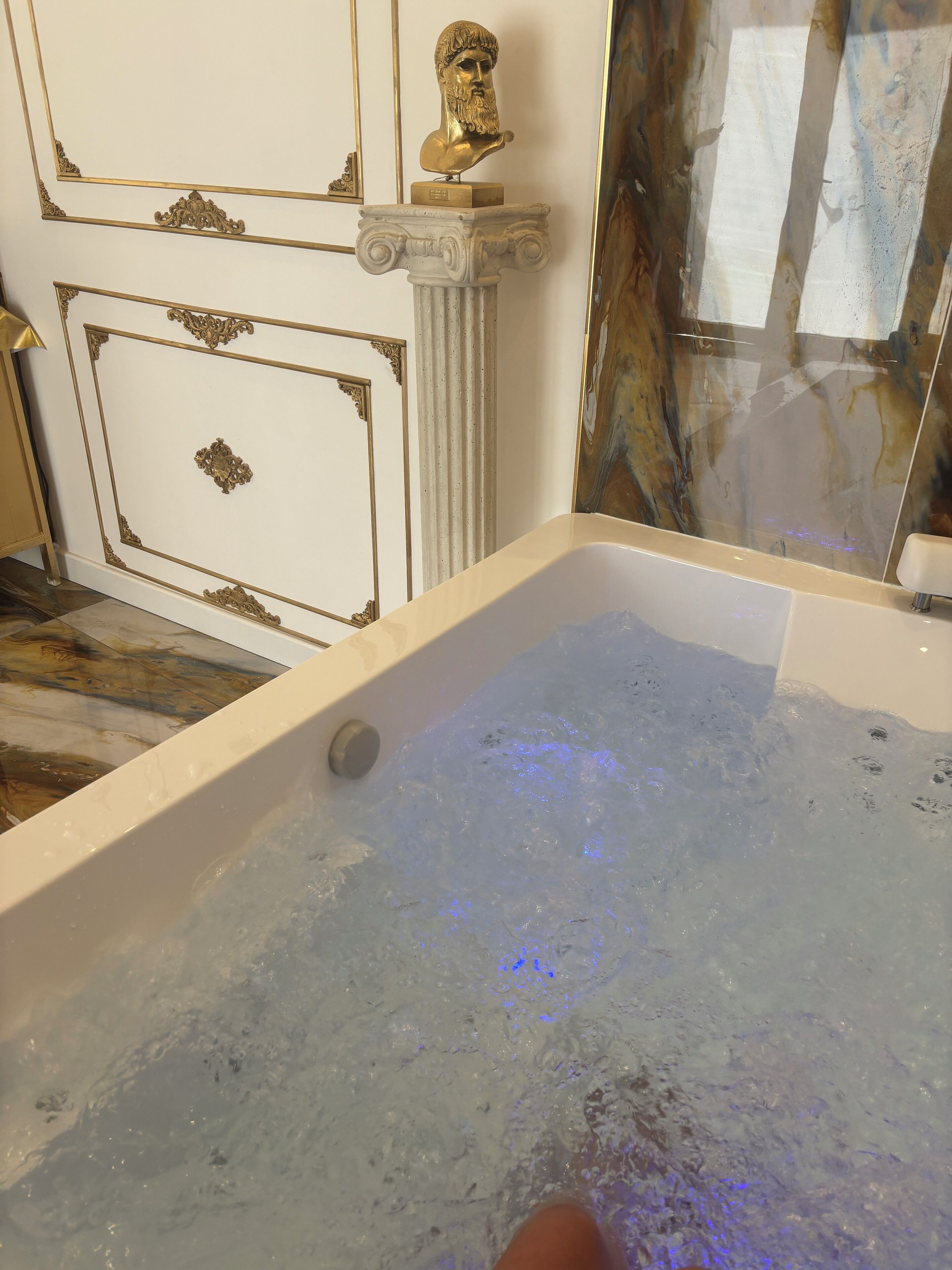 Eine Badewanne mit fließendem Wasser, ein goldener Büste auf einem Sockel und eine Marmorwand.