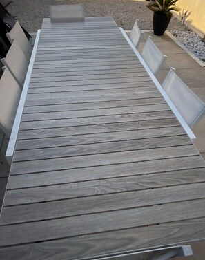 Een lange, smalle houten tafel met witte stoelen aan weerszijden, op een betonnen terras met een potplant op de achtergrond.