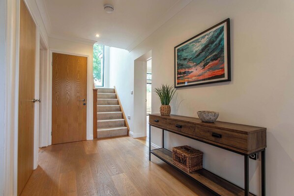 A hallway with wooden flooring, a staircase, a console table with a bowl and plant, and a framed painting on the wall.