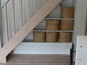 Un escalier avec une rampe en bois et un mur blanc orné de panneaux horizontaux. Trois étagères, sur lesquelles sont posés des paniers en osier, sont fixées au mur.
