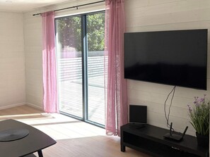 Un salon moderne avec un grand téléviseur à écran plat fixé au mur, une porte-fenêtre coulissante donnant sur un espace extérieur et une petite table noire avec une télécommande.