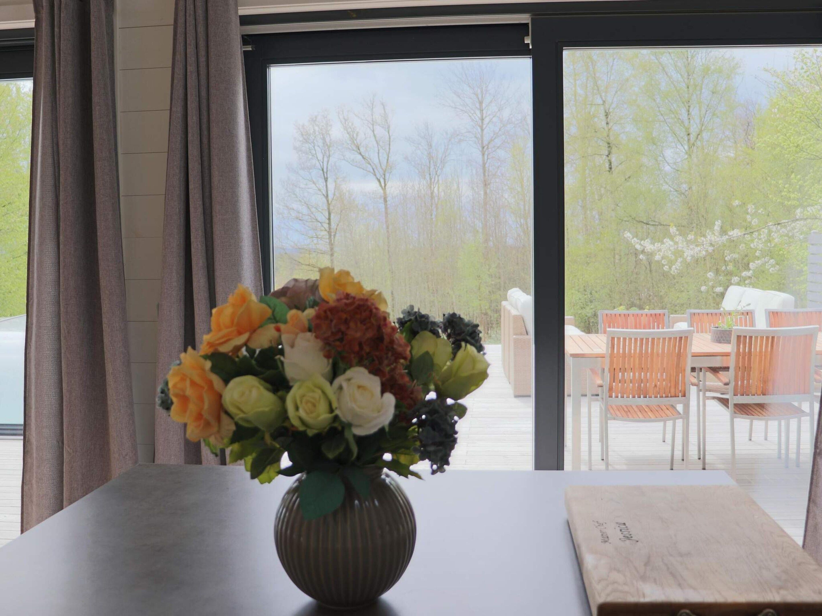 A room with a large window, a vase of flowers on a table, and a view of an outdoor deck with chairs and a table.
