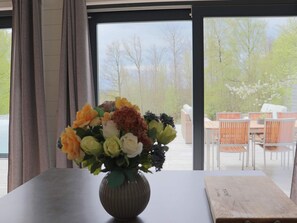 Une pièce dotée d’une grande fenêtre, d’un vase rempli de fleurs posé sur une table, et offrant une vue sur une terrasse extérieure avec des chaises et une table.
