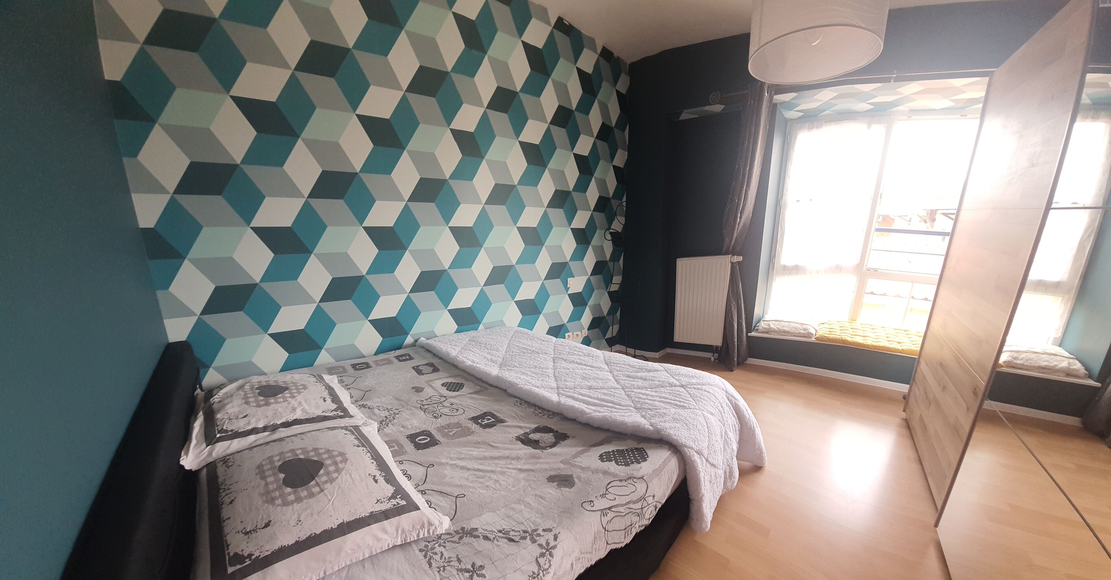 A bedroom with a geometric-patterned wall, a bed with patterned bedding, a radiator, and a window with curtains.