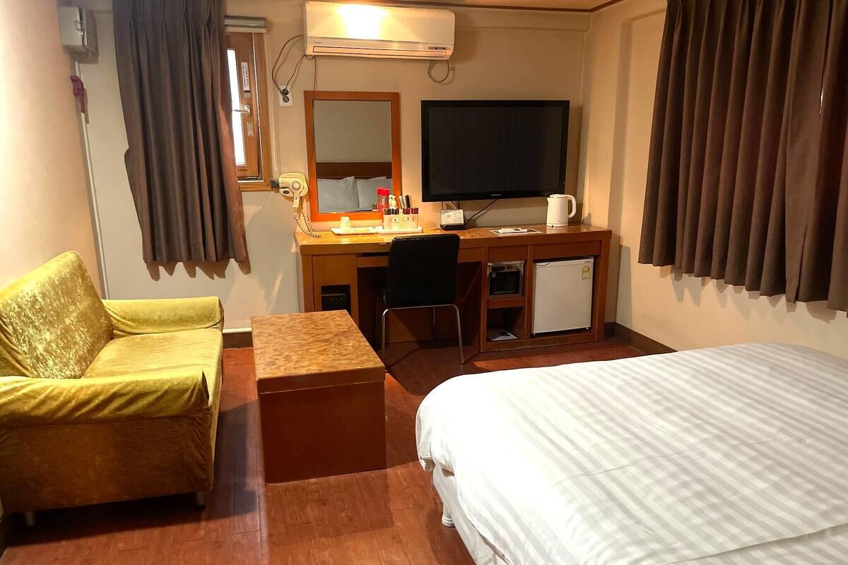 A hotel room with a bed, a desk with a television, a chair, a small refrigerator, and a yellow armchair.