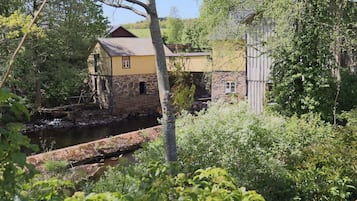 Un edificio rústico con base de piedra y fachada amarilla, rodeado de abundante vegetación y un pequeño canal de agua.