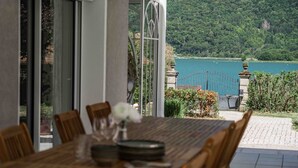 A dining table with chairs, a vase with flowers, and a view of a lake and forest. - Villa Mariette, large charming villa with lake view, swimming pool and large garden (Doussard)