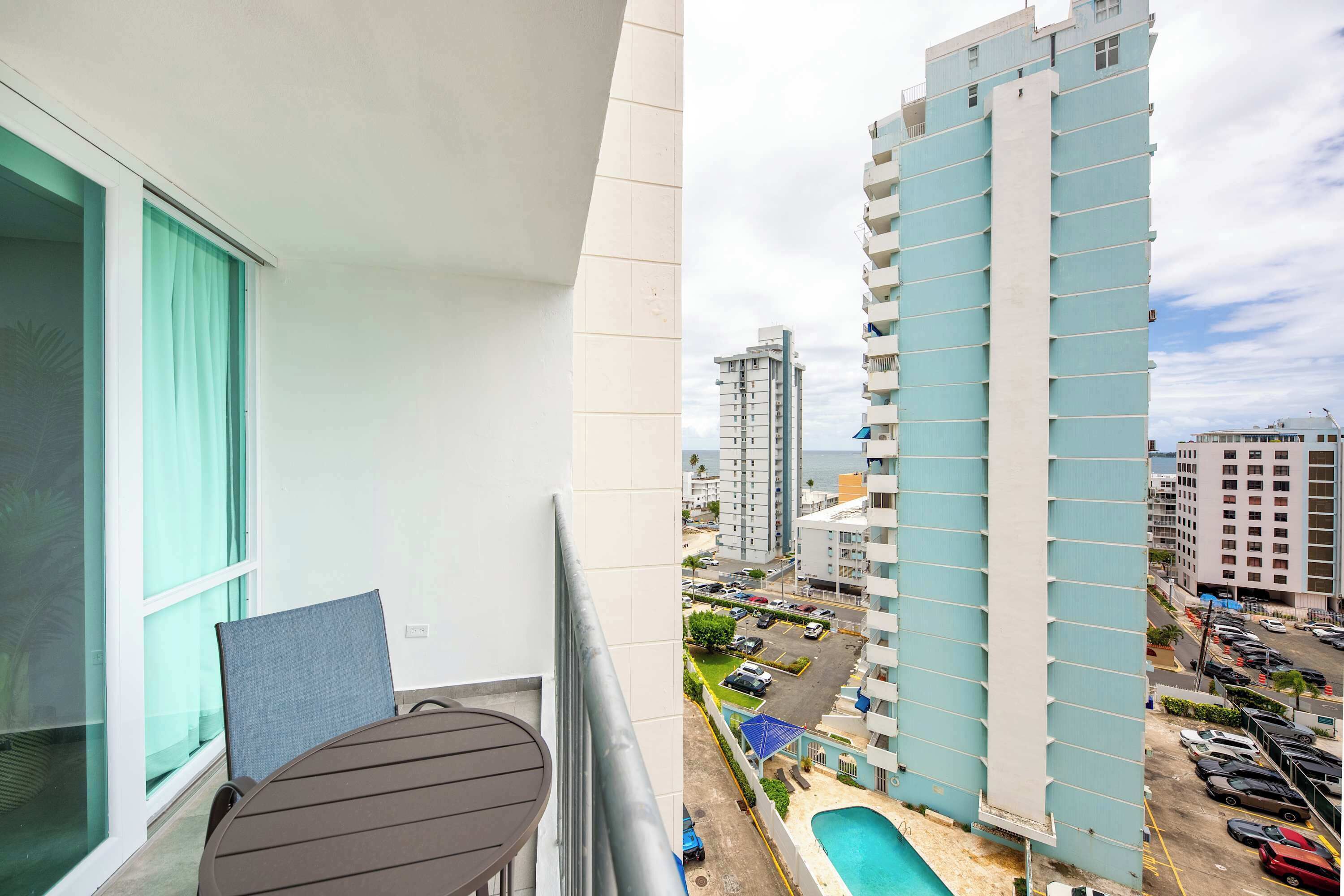 A balcony with a table and chairs, overlooking a parking lot and a tall building with a pool.