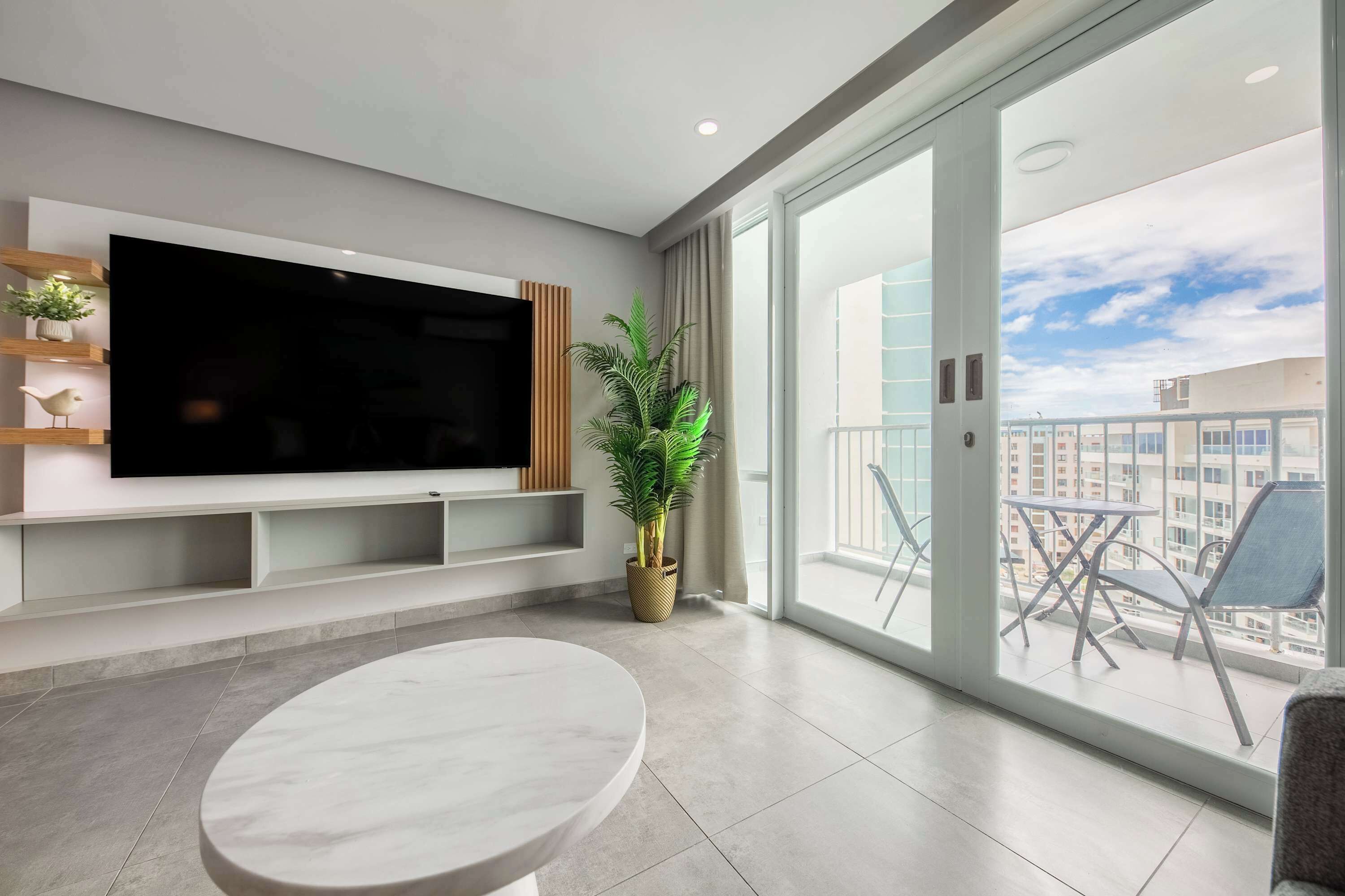 Een moderne woonkamer met een grote flatscreen televisie, een ronde marmeren tafel, een potplant en een balkon met buitenmeubilair.