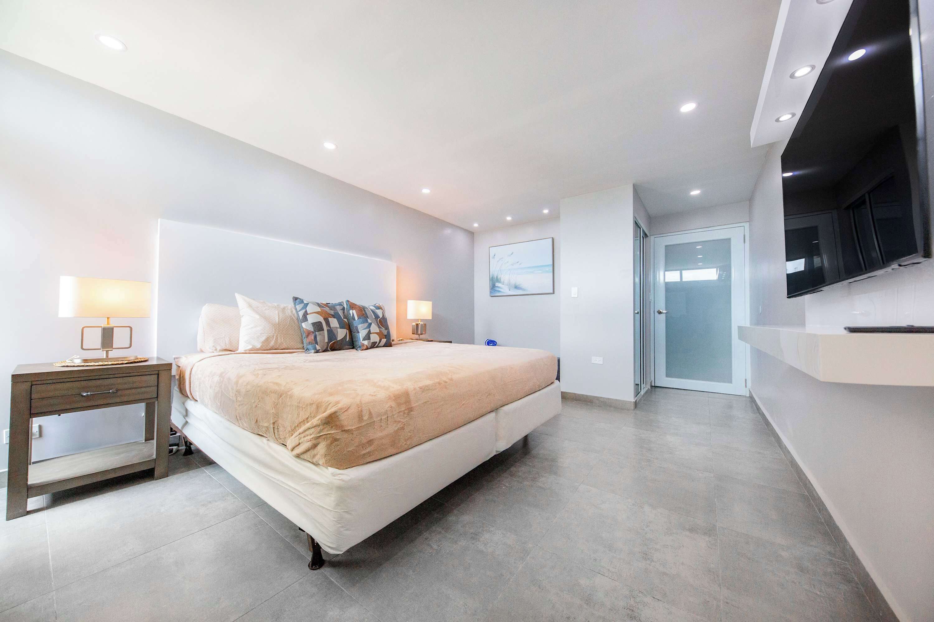 Un dormitorio moderno con una cama grande, mesitas de noche, un televisor y una puerta que da a otra habitación.