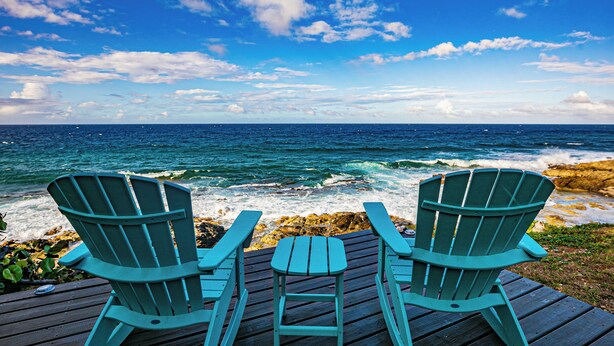 Twee groene Adirondack-stoelen op een houten terras met uitzicht op een rotsachtige kustlijn, waar golven tegen de rotsen slaan, onder een heldere blauwe lucht.