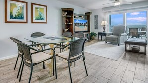A spacious living room with a glass-top dining table surrounded by wicker chairs, a television on a wooden cabinet, and a view of the ocean through large windows.