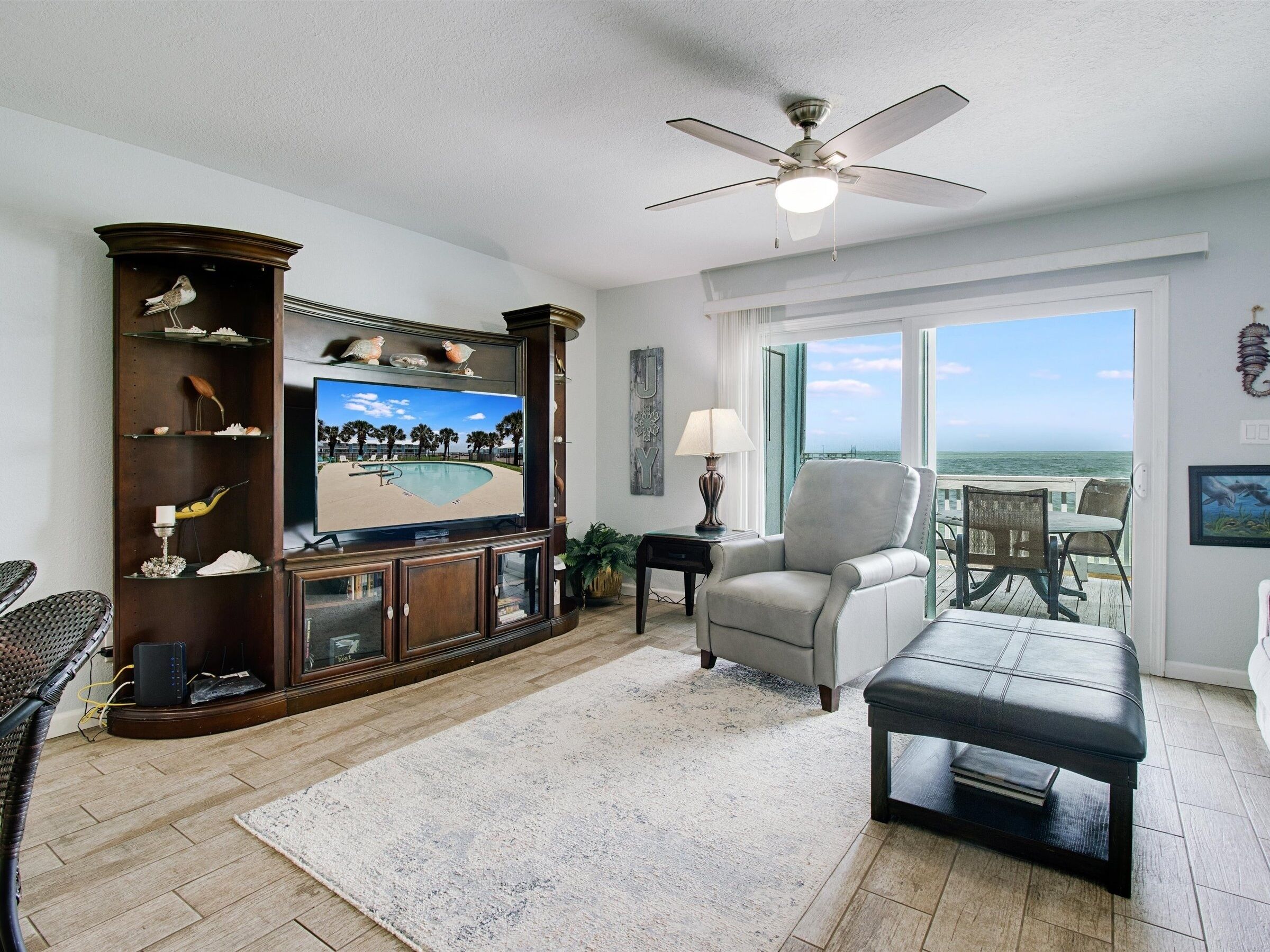 A living room with a large flat-screen TV, a ceiling fan, a view of the ocean through large windows, and a balcony with outdoor furniture.