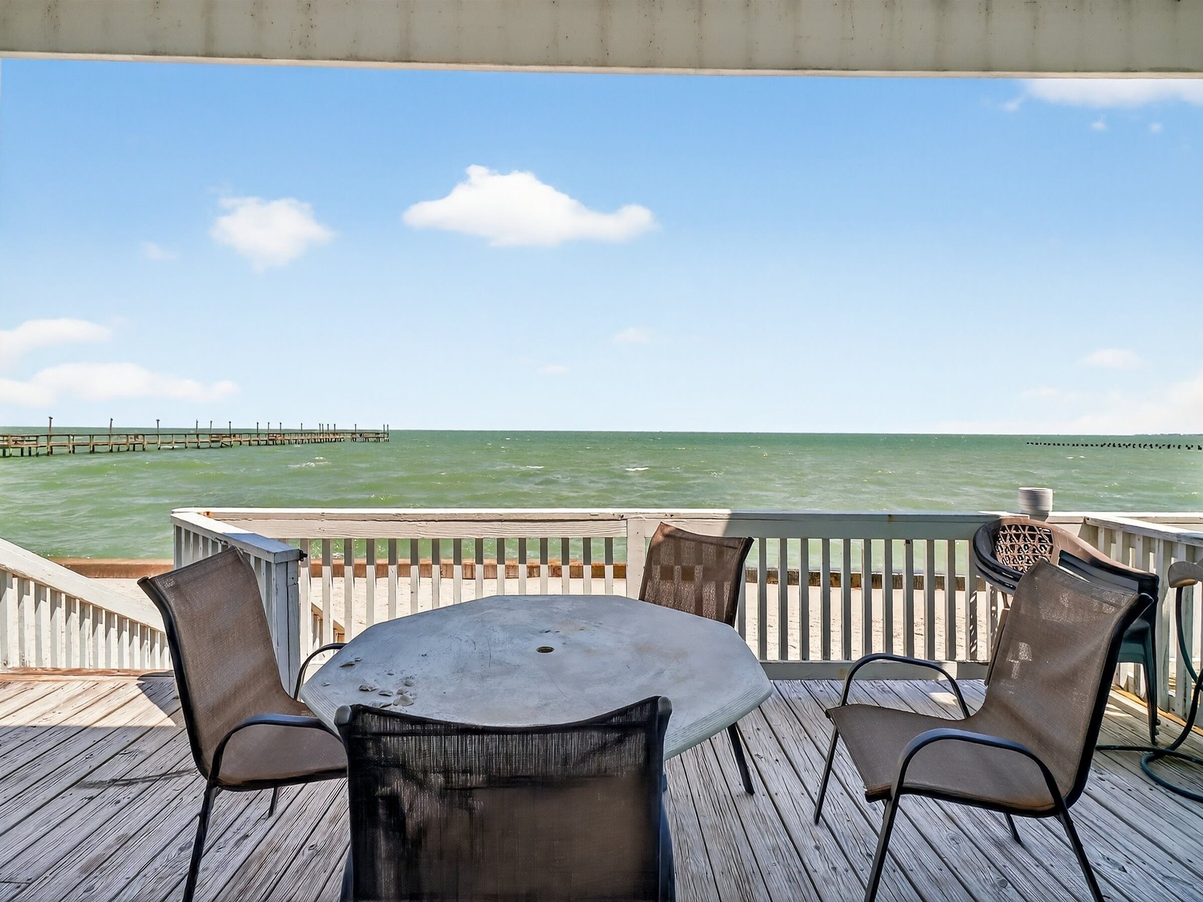 A deck with a table and chairs overlooking the ocean, a pier in the distance, and a clear sky.