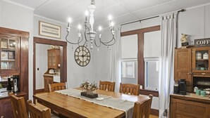 A dining area with a wooden table, chairs, a chandelier, a wall clock, and a kitchen area with cabinets and a coffee station. - 1929 Craftsman Charmer 2 Bedroom Home (Fredericksburg)