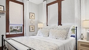 A bedroom with a four-poster bed, a nightstand with a lamp, and a view of a street through the window. - 1929 Craftsman Charmer 2 Bedroom Home (Fredericksburg)