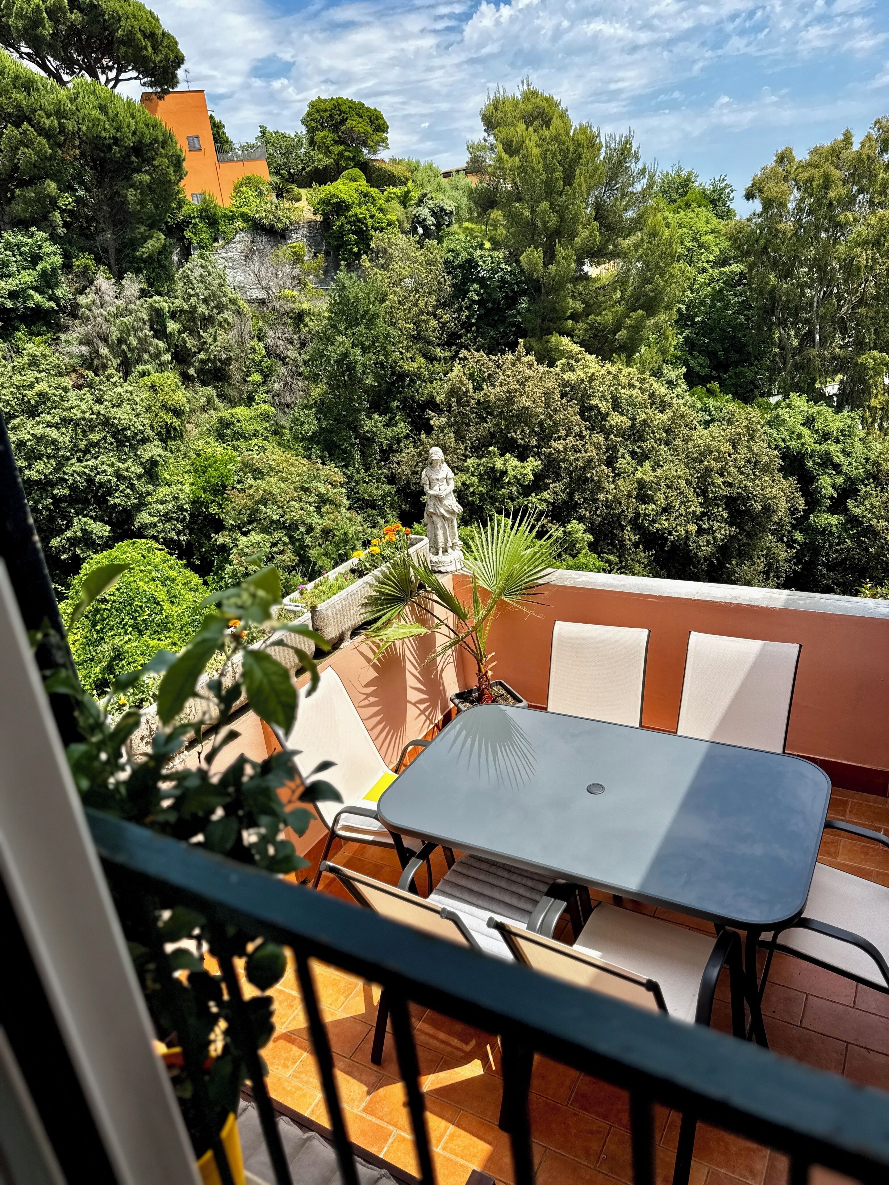 Un balcon agrémenté d’une table et de chaises, entouré d’une végétation luxuriante et orné d’une statue.