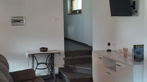 Una sala moderna con un sofá, una mesa con mantel estampado, techo de madera, una escalera, un televisor montado en la pared y una pequeña mesa con un adorno.