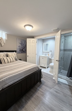 A bedroom with a bed, a bedside table, a dresser, a mirror, and a shower area. - Fabulous 2-bedroom apartment with AC in charming Whitby (Whitby)