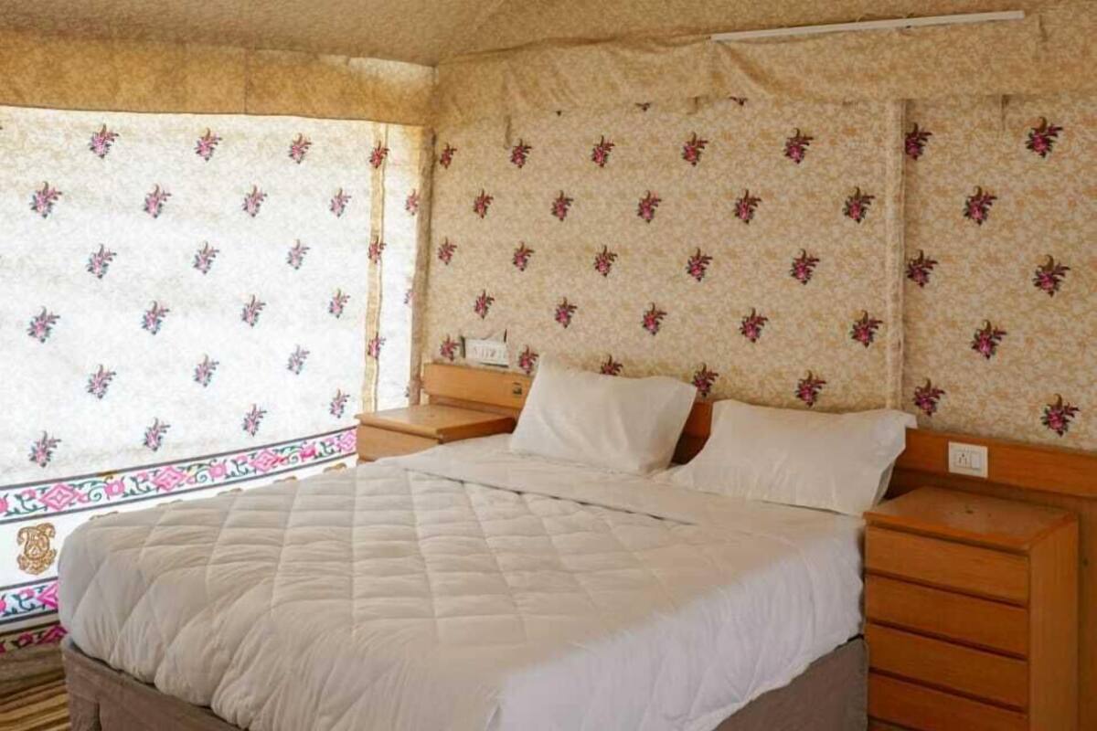 Quarto com cama, mesa de cabeceira, cômoda de madeira, ventilador de teto e papel de parede com padrão floral.
