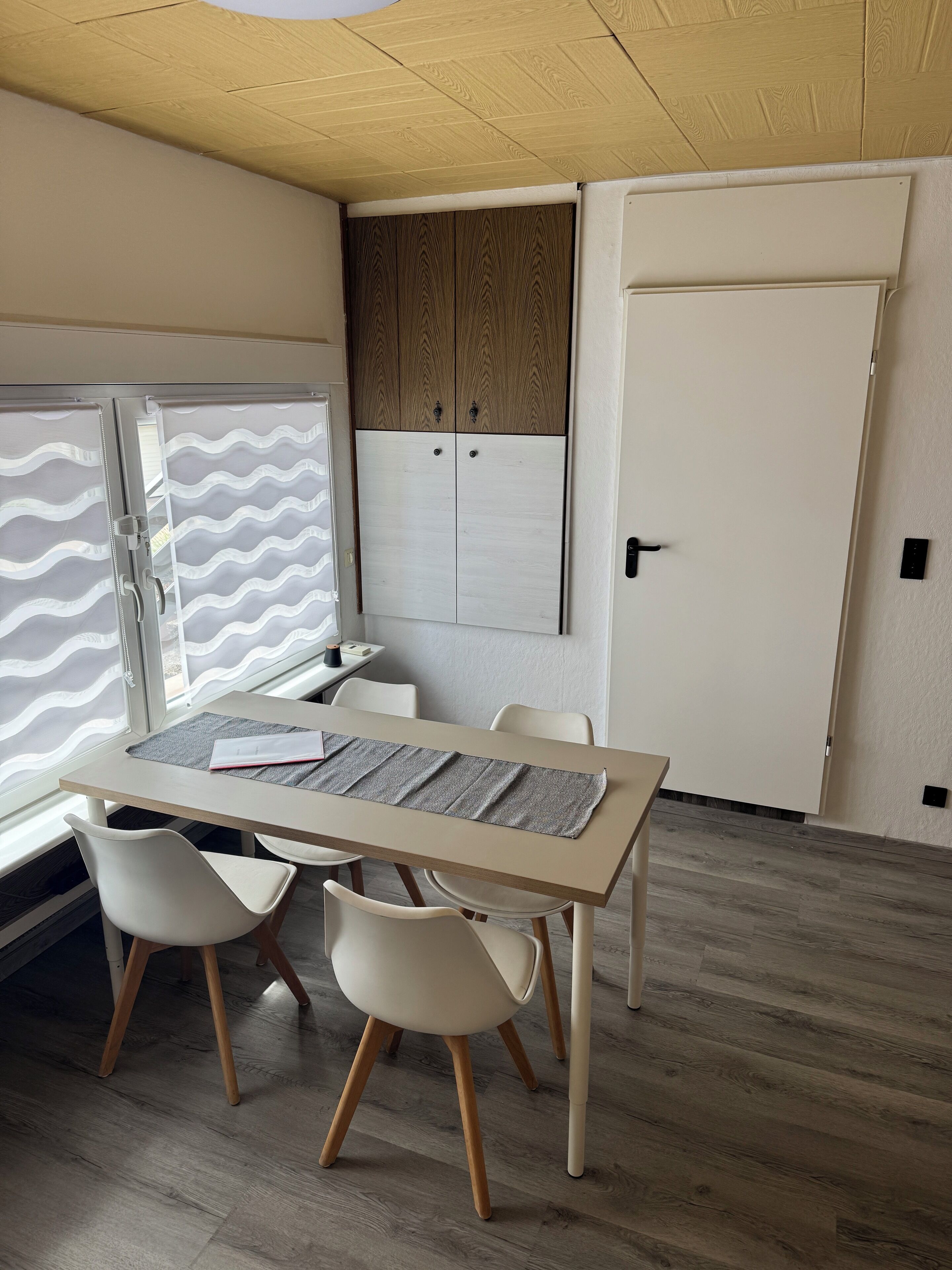 Ein Zimmer mit Holzboden, einem Tisch mit zwei Stühlen, einem Fenster mit wellenförmigen Vorhängen und einer Holztür.