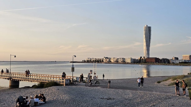 A riverside promenade with people walking, sitting, and cycling. A distinctive tower-like building is visible in the background. - 2 Person Comfortable Holiday Park Home in Testby (Malmö)
