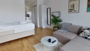 A modern, minimalist living room with a white sofa, a round coffee table, and a grey couch. There is a potted plant and a framed picture of a building on the wall. - 2 Person Comfortable Holiday Park Home in Testby (Malmö)