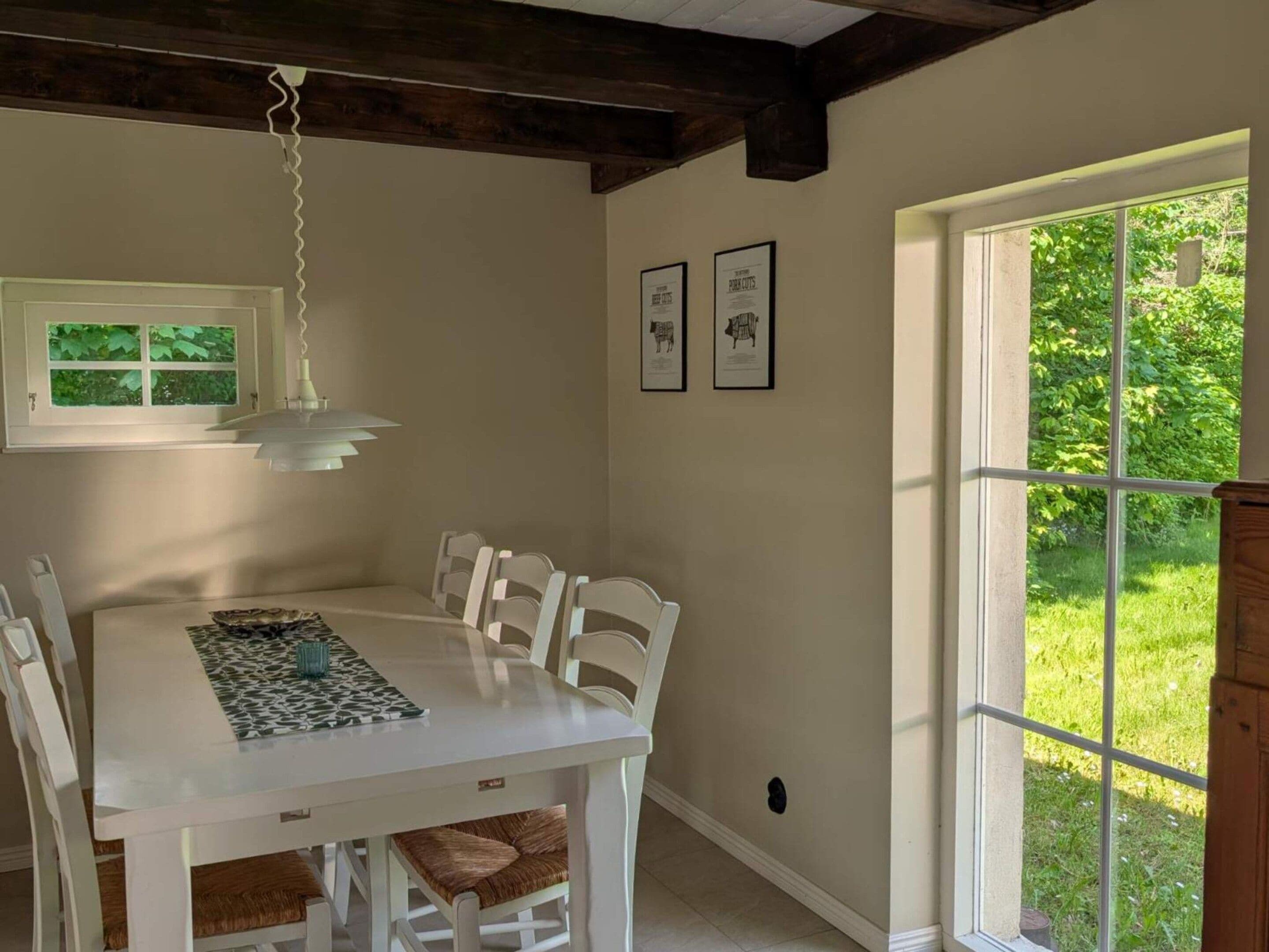 A dining area with a white table and chairs, a hanging light fixture, framed pictures on the wall, and a window with a view of greenery.