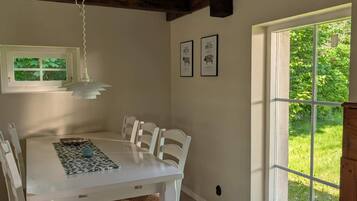 A dining area with a white table and chairs, a hanging light fixture, framed pictures on the wall, and a window with a view of greenery.