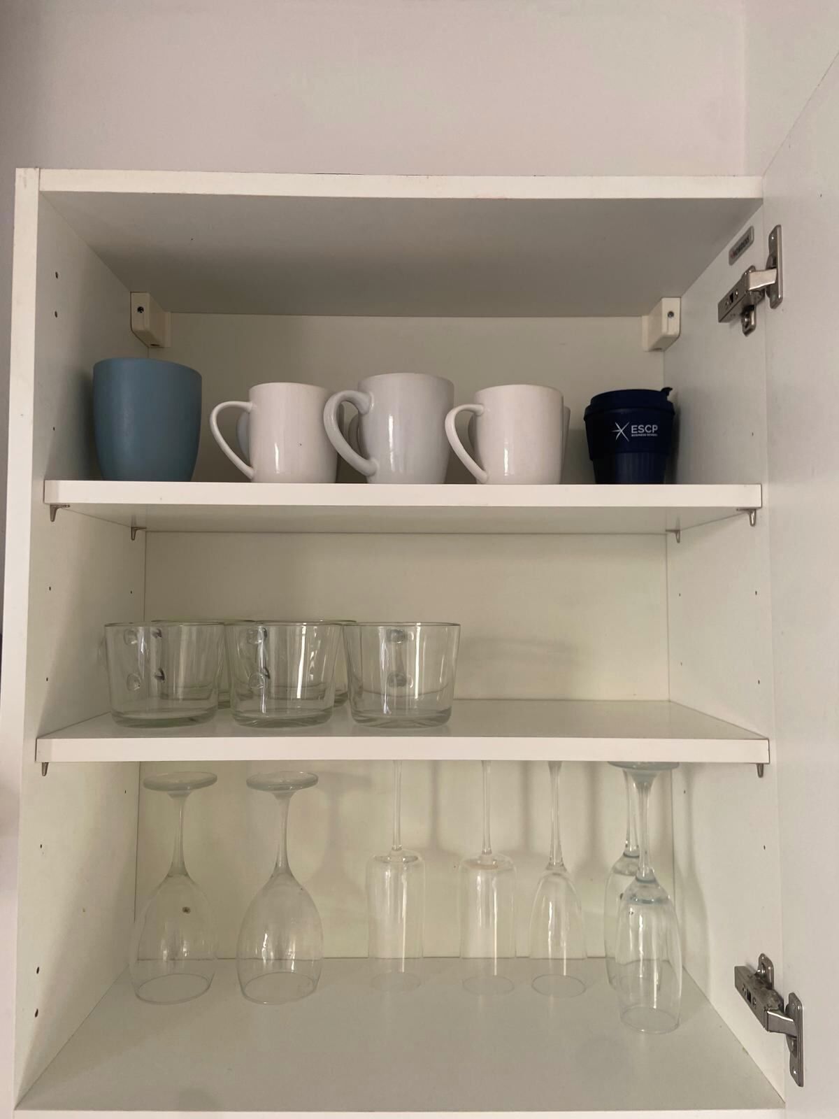 Un gabinete blanco con tres estantes. El estante superior sostiene cuatro tazas blancas y una taza azul. El estante central tiene cuatro vasos de vidrio transparente. El estante inferior contiene seis copas de vino.