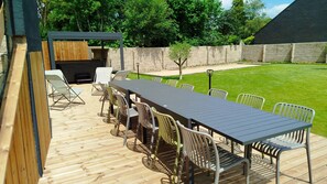 Une terrasse extérieure avec une longue table et des chaises, un barbecue et un petit arbre.