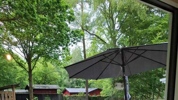 Une terrasse en bois, agrémentée de mobilier d’extérieur et d’un grand parasol, entourée d’une végétation luxuriante et d’une clôture en bois.