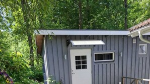 Un petit abri en bois d’un seul étage, avec une porte blanche et une fenêtre, entouré d’une allée gravillonnée et d’une végétation luxuriante.