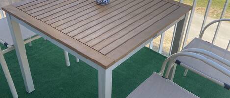 Un balcón con mesa de madera y sillas blancas, un pequeño recipiente decorativo y vista a un área residencial.