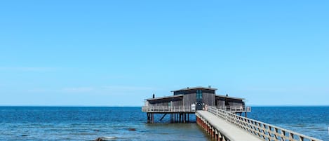 Un muelle de madera que conduce a un edificio de techo plano, ubicado en una playa arenosa con un mar tranquilo al fondo.