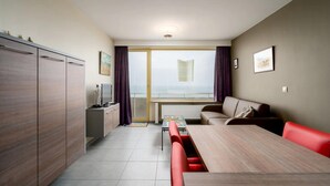 Moderni hotellihuone, jossa on ruokailutila, sohva, televisio ja suuri ikkuna, jossa on verhot.