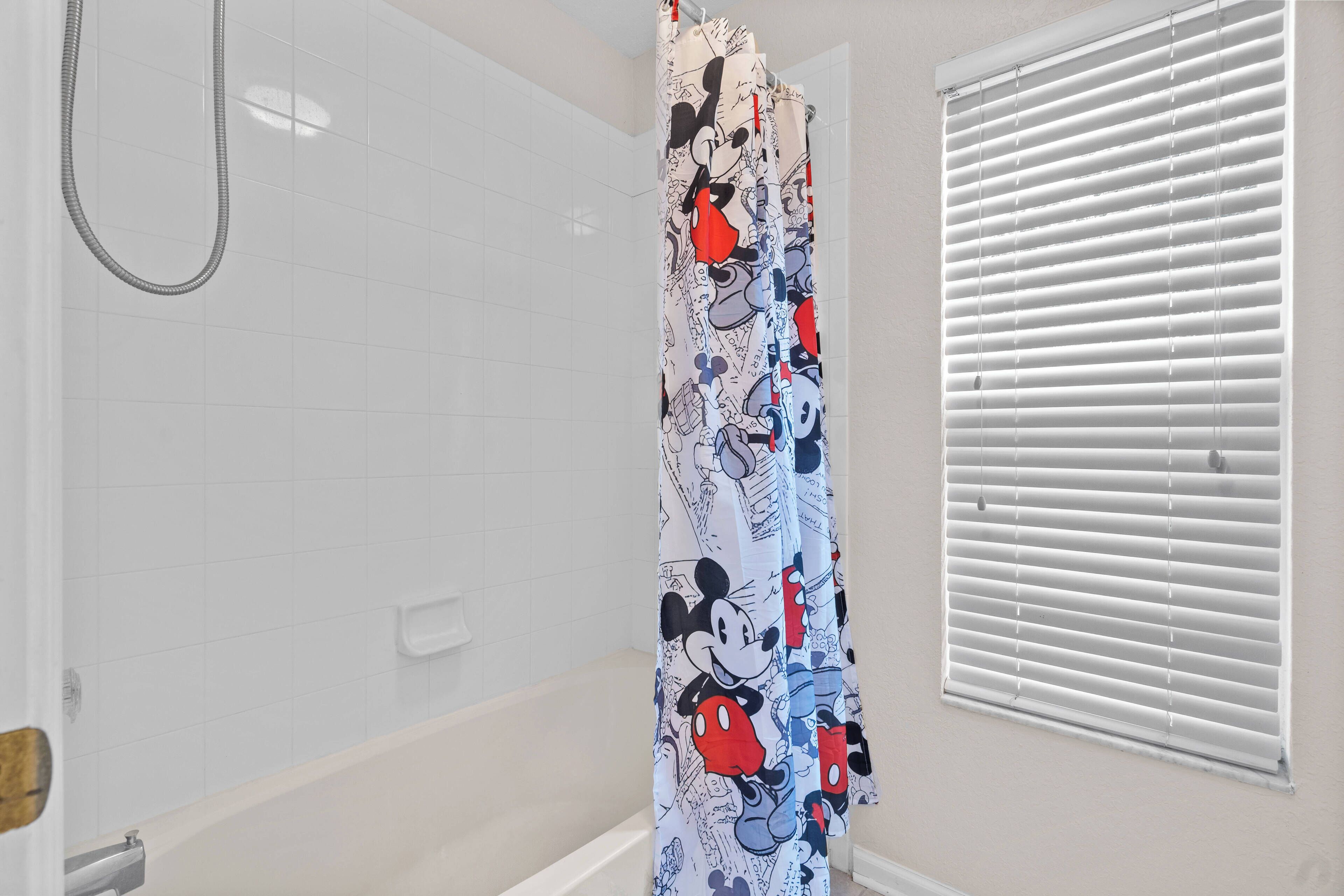 Une salle de bain avec un rideau de douche orné de personnages de dessins animés, une pomme de douche et une fenêtre avec des stores.