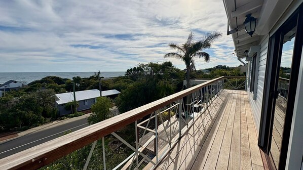 A wooden deck with a metal railing overlooking a road and a body of water. - Peppy Beach House – Stunning Ocean Views & Relaxed Coastal Escape (Peppermint Grove Beach)