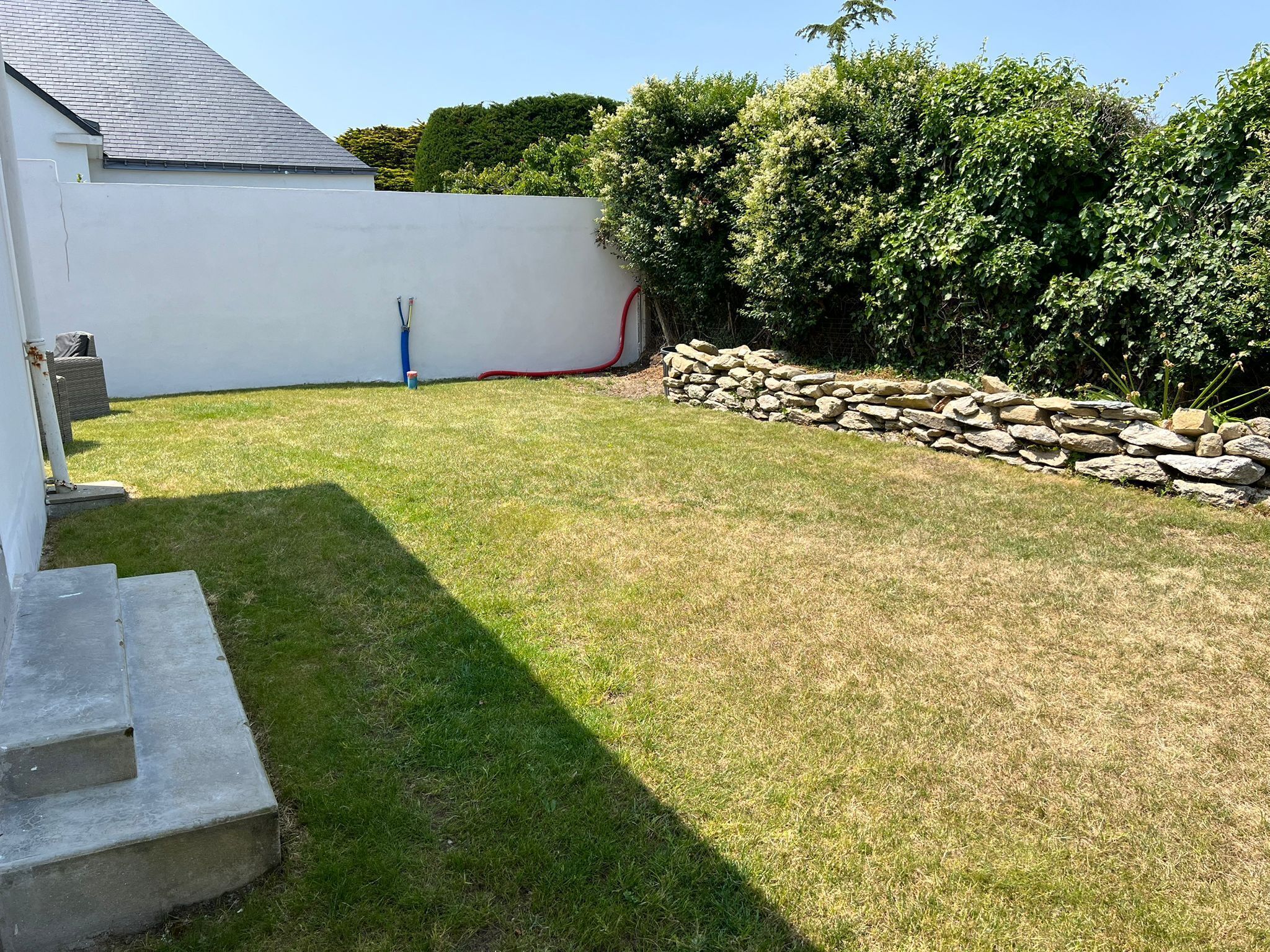 A backyard with a stone wall, a grassy lawn, and a white building with a grey roof.