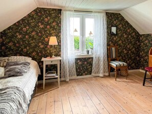 Un dormitorio con techo inclinado, papel tapiz floral, una cama con manta blanca, una mesita de noche con lámpara, una silla con cojín y una ventana con cortinas de encaje.