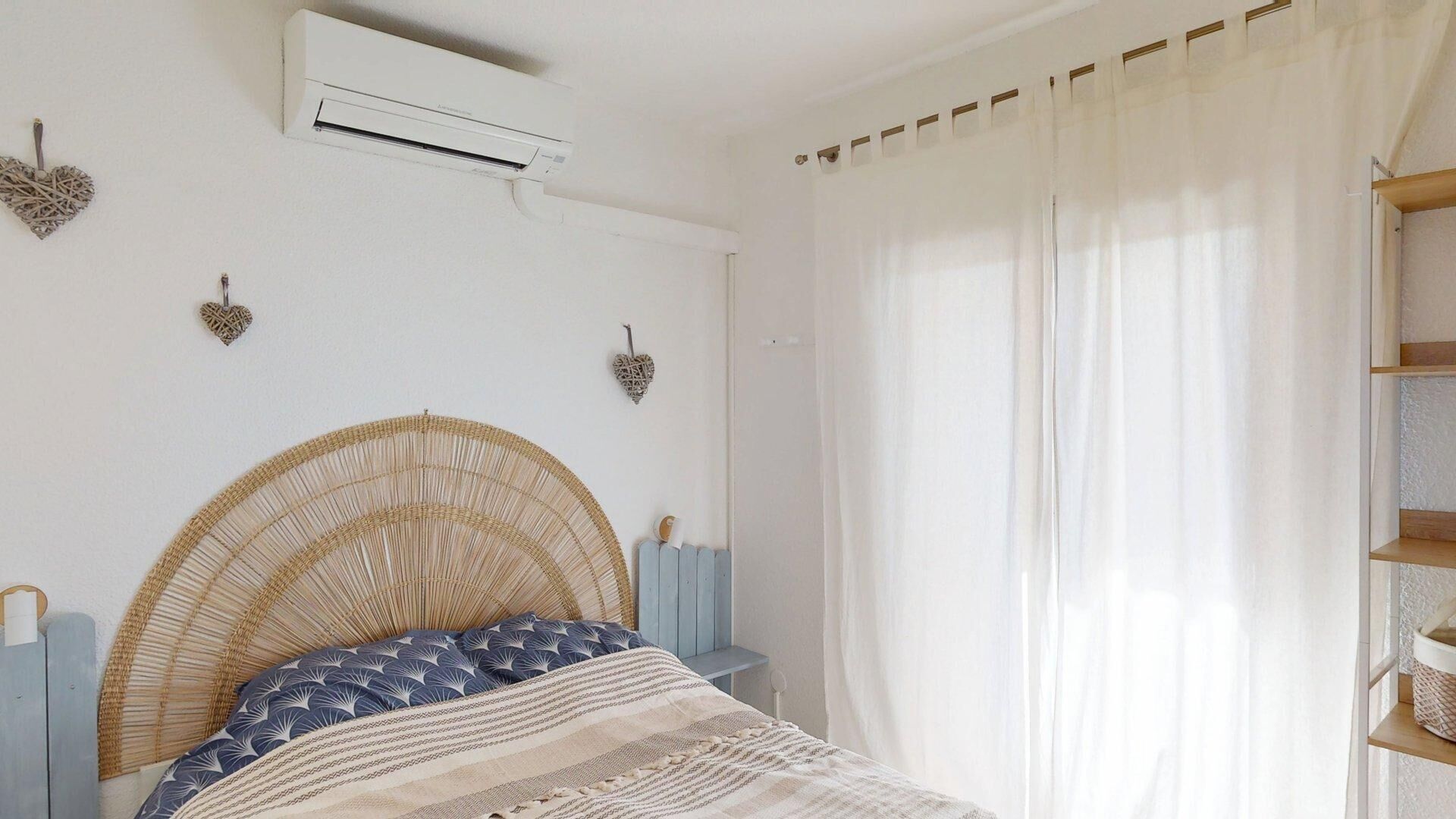 A bedroom with a bed, an air conditioner, a window with sheer curtains, and decorative heart-shaped wall hangings.